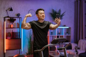 Young asian man showing bicep while looking at smartphone during exercising on treadmill.